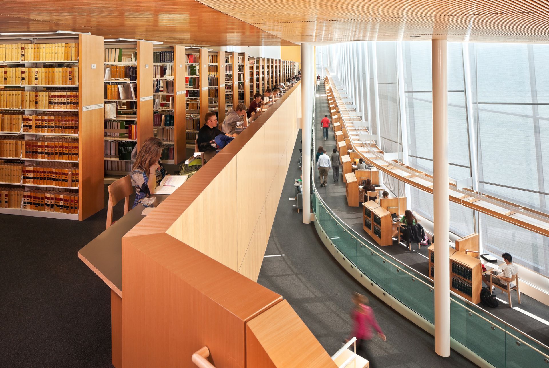 Multilevel library interior with rows of shelves, tables, and wall of windows.